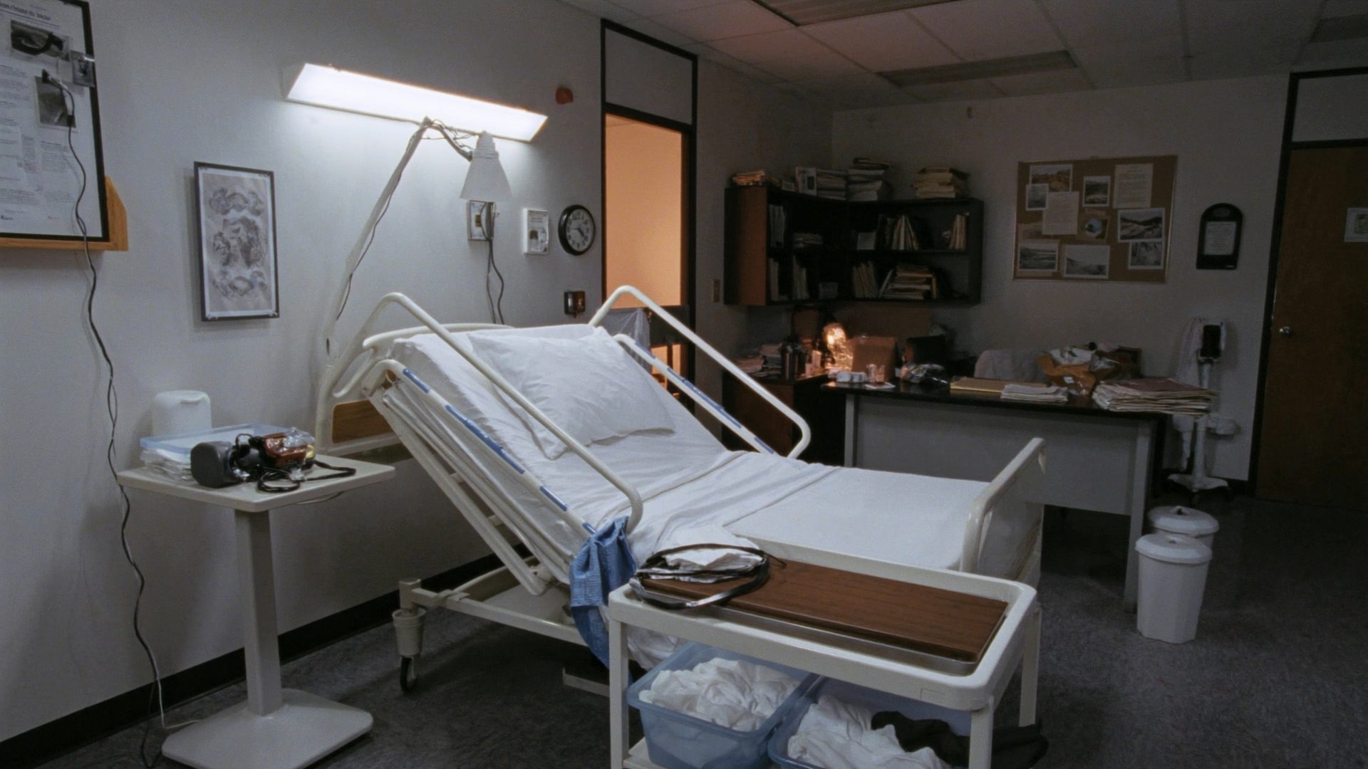 A nurse aide skills lab in an evening classroom: a hospital bed made up with white linens, a blood pressure cuff and stethoscope on the bedside table, a gait belt and transfer board, and a rolling cart with a basin and towels.